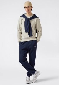 Sudadera gris clara con detalles en azul marino, pantalones deportivos azul marino y gorro de punto beige. Presenta logotipos en el pecho y en los pantalones. Zapatillas texturizadas.