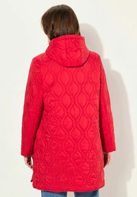 Red quilted coat with a hood, featuring a wave pattern and side slits for movement, paired with blue jeans.