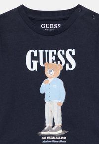 Κοντομάνικο μπλουζάκι σε ναυτικό μπλε χρώμα με καρτούν αρκούδα που φοράει ανοιχτό μπλε μπουφάν και ριγέ παντελόνι, με το λογότυπο "GUESS" να είναι ευδιάκριτο πάνω από την εικόνα.