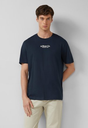 Jonge man met kort bruin haar draagt een navyblauw T-shirt met de tekst "s.Oliver Co. Established 1969" en beige broek tegen een effen grijze muur.