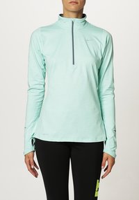 Kobieta ubrana w jasnoturkusową sportową bluzę z długim rękawem i półzamkiem z przodu oraz czarne legginsy z żółtym wzorem na lewej uda.