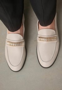 Vaaleanharmaat nahka loaferit, joissa on koristeellinen kultainen yksityiskohta etuosassa, pyöreä kärki ja musta kumipohja. Sopivat arkikäyttöön.