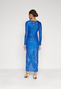 Μακρύ μπλε φόρεμα με παγιέτες, μακριά μανίκια, εφαρμοστή σιλουέτα, στρογγυλή λαιμόκοψη και κεντρικό φερμουάρ στην πλάτη. Περιλαμβάνει λευκά πέδιλα με straps.