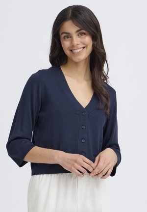 Giovane donna sorridente, indossa un cardigan blu navy con bottoni e maniche a tre quarti e una gonna bianca, in piedi davanti a uno sfondo semplice.