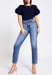 Mörkblå topp med volangärmar, ljusblå högmidjade jeans med fransiga fållar, kombinerade med beige remsandaler. Enkel, avslappnad outfit med tydliga texturer.