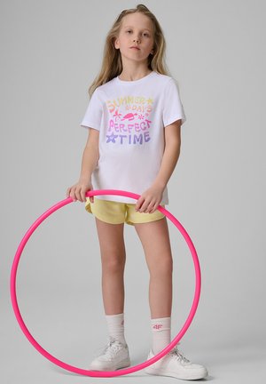 Dziewczyna w białej koszulce "Summer Days Perfect Time" i żółtych szortach trzymająca różowy hula-hop na szarym tle.