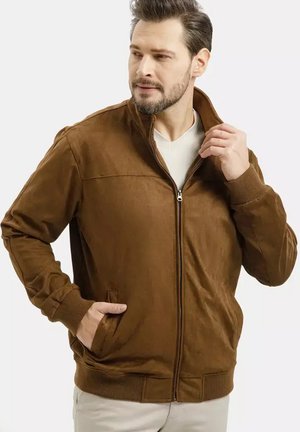 Mężczyzna z brodą, ubrany w brązową kurtkę na zamek błyskawiczny na białą koszulę, jedną ręką w kieszeni, poprawia kołnierz, patrząc na bok.