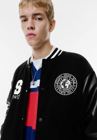 Sort varsity-jakke med hvide striber, broderet logo og læderærmer. Lagd med en flerfarvet stribet skjorte under.