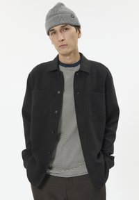 Sort knapjakke med to forlommer, båret over en marineblå og hvid stribet skjorte. Grå strikket beanie fuldender looket.