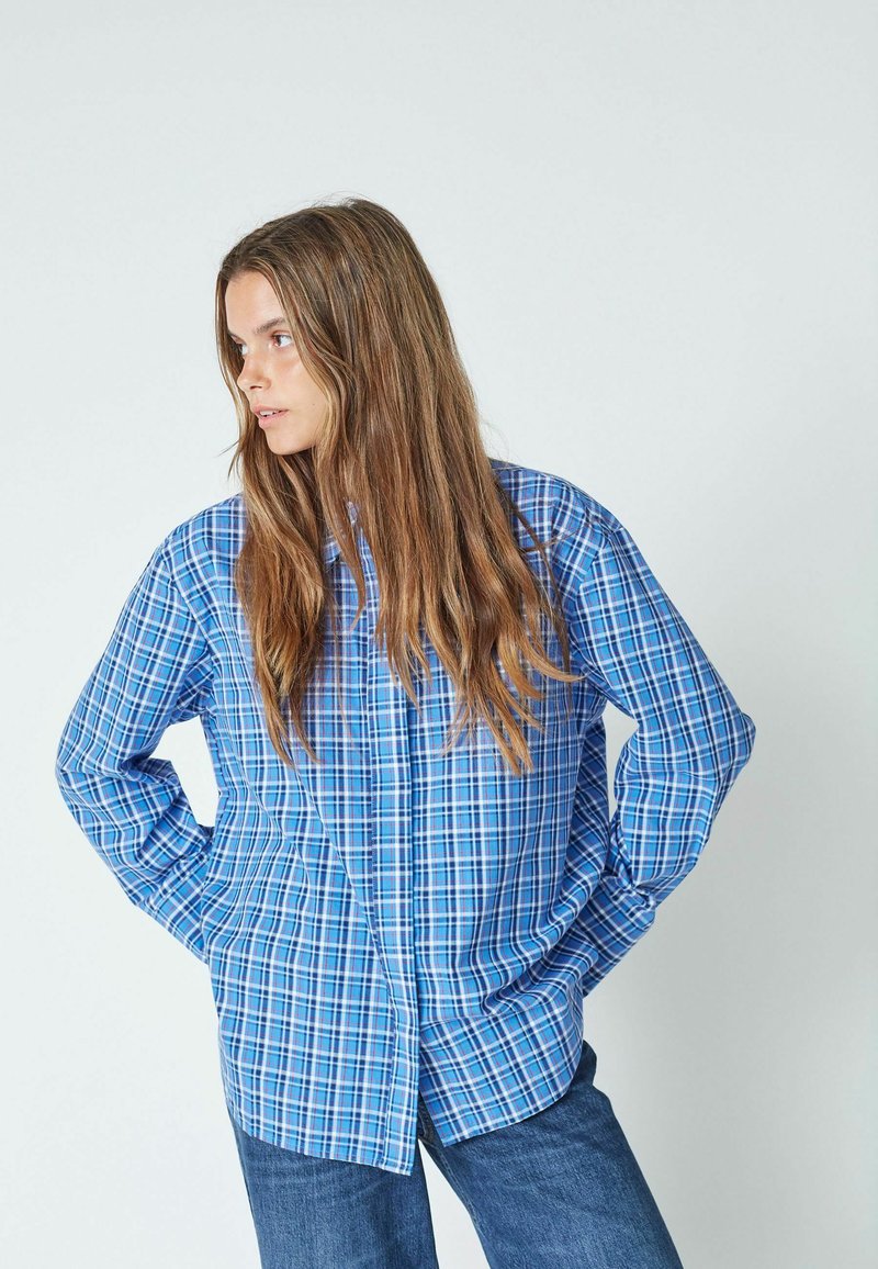 Modra karirana srajca s gumbi, izdelana iz tkanine s sproščenim krojem. Ima dolge rokave in rahlo ukrivljen rob, v kombinaciji s temnimi kavbojkami.