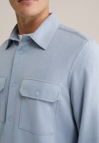 Cămașă albă-albăstruie, dintr-un material moale, cu guler ascuțit, două buzunare frontale cu clapete și detalii subtile de cusătură.