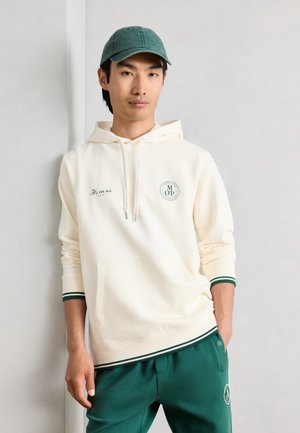 Νέος άνδρας που φοράει μια κρεμ-φούτερ με πράσινες λεπτομέρειες και ταιριαστό πράσινο παντελόνι και καπέλο, στέκεται μπροστά σε έναν ανοιχτό τοίχο.