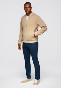 Beżowa kurtka na zamek z miękkiego materiału, z prążkowanym kołnierzem i mankietami, z bocznymi kieszeniami. Połączona z ciemnymi dżinsami i jasnobeżowymi trampkami.