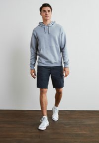 Grauer Hoodie mit Fronttasche, kombiniert mit dunkelblauen Shorts. Das Modell trägt weiße High-Top-Sneaker auf einem Holzboden vor einem schlichten Hintergrund.