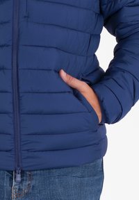 Persoon in een blauwe gewatteerde jas met zijzakken, hand gedeeltelijk in de rechterzak, gecombineerd met blauwe denim jeans.