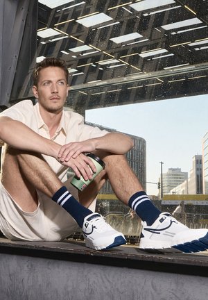 Weiße und marineblaue Sportschuhe mit strukturierten Außensohlen. Das Modell trägt marineblaue gestreifte Socken und ein helles kurzärmliges Outfit und hält eine grüne Tasse.