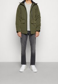 Giacca a vento verde oliva con cappuccio, dotata di due tasche frontali e una tasca sul petto con zipper, abbinata a una t-shirt grigia e jeans grigio scuro. Sneakers bianche.