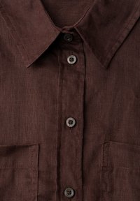 Gros plan sur le devant d'une chemise marron foncé à col avec trois boutons visibles et deux poches poitrine, montrant la texture du tissu et les détails de couture.