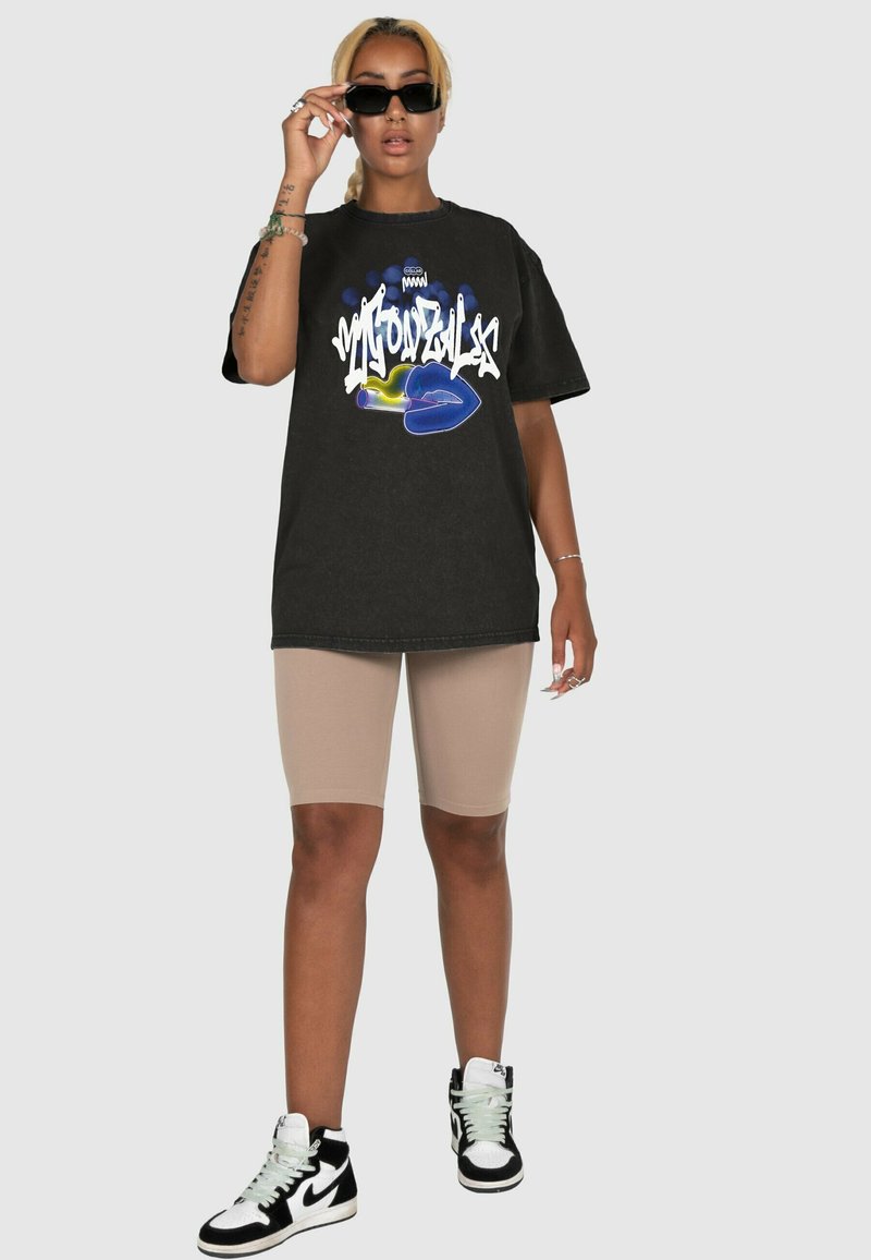 Fekete, nagyméretű póló grafikus nyomattal, kék ajkakkal és fehér szöveggel. Bézs kerékpáros rövidnadrág és fekete magasszárú sportcipő egészíti ki a szettet.