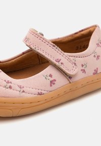 Rozā zamša kurpe ar ziedu rakstu, ar Velcro siksniņu, noapaļota purngala un bēša gumijas zole ar diskrētu diegu. 