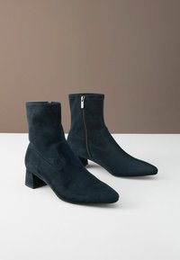 Mörkblå mockaskor med ankelhöjd, spetsig tå och blockklack. Har en sidodragkedja och en slät yta med minimala detaljer.