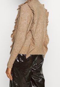 Pull en tricot beige avec des bords volants, présentant une texture douce. Assorti à un pantalon en vinyle noir brillant. Vue de dos montrée.