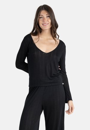 Jeune femme aux longs cheveux bruns portant un haut noir à manches longues et un pantalon noir assorti, souriante avec une main derrière le dos.