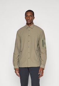 Ελαιόχρωμο μπουφάν φτιαγμένο από υφασμάτινο, υφή, με κουμπιά που ασφαλίζουν, τσέπη στο στήθος και ρυθμιζόμενο στρίφωμα. Περιλαμβάνει μία θηλή στο μανίκι.