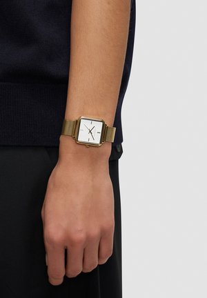 Vierkante gouden horloge met een witte wijzerplaat, zwarte uurmarkeringen en een gouden metalen mesh band, gedragen aan een hand tegen een donkere kledingachtergrond.