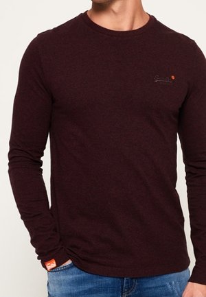 Homme portant une chemise ajustée à manches longues de couleur bordeaux foncé avec un petit logo brodé sur la poitrine et une étiquette orange au poignet, associée à un jean bleu.