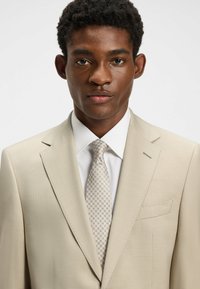 Beige blazer med en subtil tekstur, notched lapels og en lomme; parret med en hvid skjorte og et let mønstret slips.