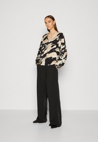 Blusa a maniche lunghe in beige, nero e crema con motivo astratto, abbinata a pantaloni neri larghi. Finitura in raso su entrambi i capi.