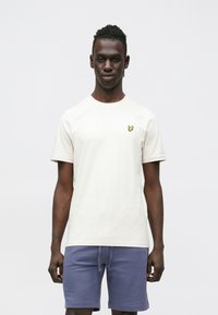 Krém színű rövid ujjú póló kerek nyakkivágással és egy kis sárga pillangó logóval a bal mellrészen; kék rövidnadrággal viselve.