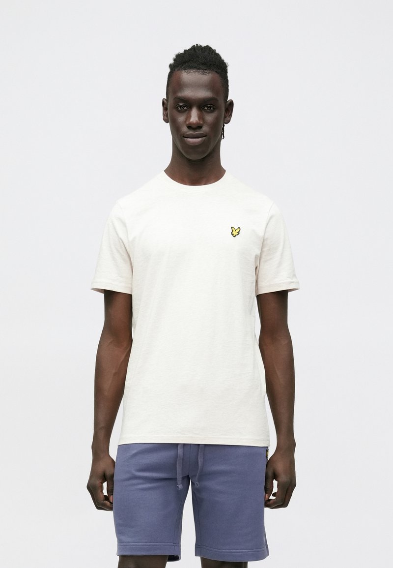 Krém színű rövid ujjú póló kerek nyakkivágással és egy kis sárga pillangó logóval a bal mellrészen; kék rövidnadrággal viselve.