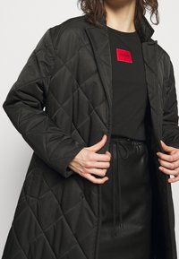 Schwarzer gesteppter langer Mantel mit Kragen, getragen über einem schwarzen Oberteil mit einem roten rechteckigen Logo. Der Mantel hat eine glatte Textur und taillierte Ärmel.