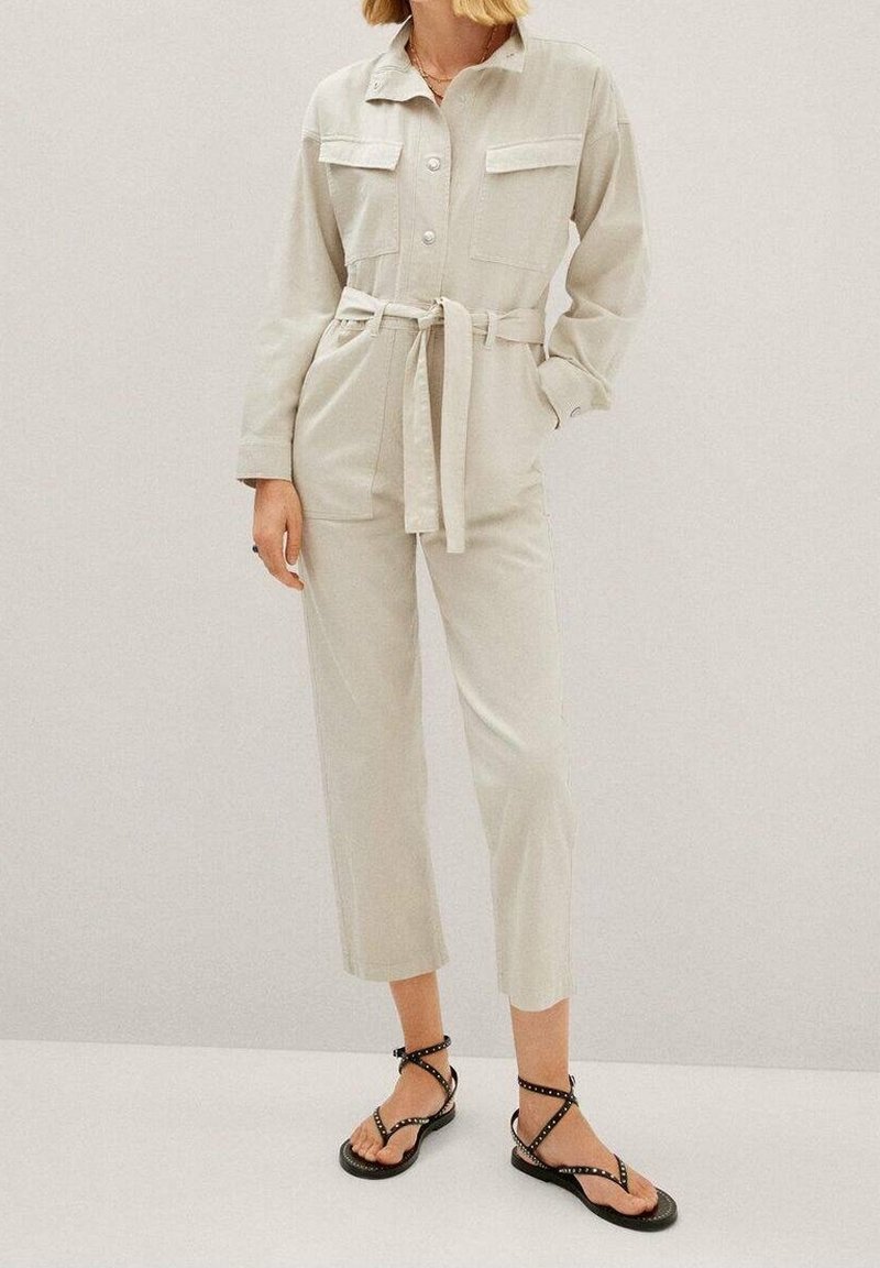 Jumpsuit beige clair à manches longues, avec un devant boutonné, deux poches poitrine et une ceinture à nouer. Associé à des sandales noires à lanières.
