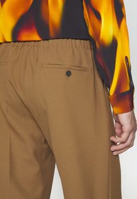 Hnědé kalhoty na míru s zadní kapsou a detaily na knoflík. Pas je elastický, což zajišťuje strukturovaný, ale pohodlný střih.