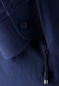 Marineblauem Stoff mit strukturiertem Finish, ausgestattet mit einer Klapptasche, schwarzem Druckverschluss und einem Kordelzug mit schwarzem und metallicfarbener Zugklemme.