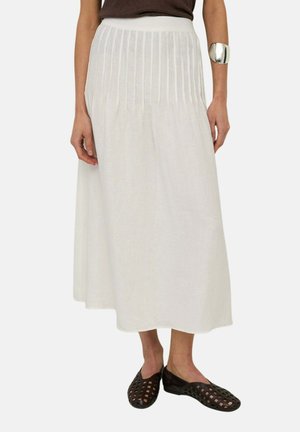 Mujer con falda midi blanca acanalada, top negro, brazalete plateado y zapatos planos negros tejidos, de pie sobre un fondo blanco.