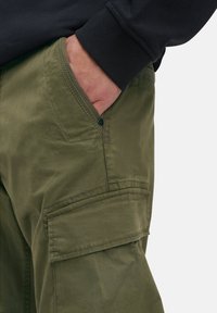 Cargo-Hosen aus olivgrünem Baumwollstoff mit glatter Textur, ausgestattet mit seitlichen Taschen und sichtbarem Knopfdetail. Der Ärmel eines schwarzen Sweatshirts ist sichtbar.