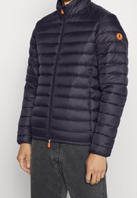 Czarna granatowa kurtka puchowa z pikowanym wzorem, wysokim kołnierzem, zapięciem na zamek błyskawiczny oraz pomarańczowymi akcentami na zamku i naszywce z logo. Lekki materiał.