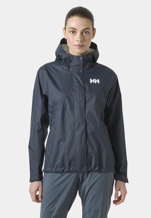 Frau trägt eine marineblaue wasserdichte Jacke mit Kapuze und weißem HH-Logo, steht mit entspannten Händen seitlich am Körper.
