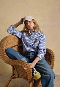 Woman in blue striped shirt, jeans, and beige cap sitting cross-legged in a wicker chair against a plain wall.