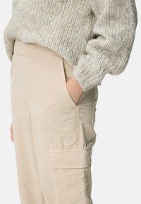 Personne portant un pantalon cargo en velours côtelé beige, une main dans la poche, et un pull en grosse maille crème à manches longues.