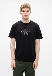 Melna īsām piedurknēm T-kreklu ar apaļu izgriezumu, centrā ar pelēko "Calvin Klein" logotipu un "ck" dizainu. Gluda kokvilnas auduma.