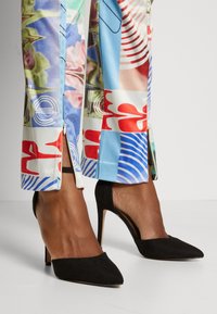 Pantalons imprimés multicolores avec des motifs bleus, rouges et floraux ; associés à des escarpins à bout pointu en daim noir avec des accents métalliques.