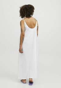Robe maxi blanche sans manches avec un dos décolleté et des détails froncés aux épaules. Le modèle porte des sandales violettes. Tissu léger et lisse.