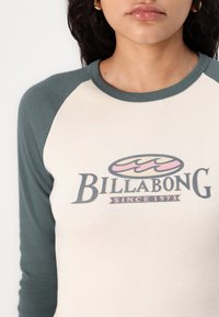 Maglietta a maniche lunghe crema e verde con scollatura rotonda, caratterizzata da un logo grafico "Billabong" e design retrò. Morbido al tatto.