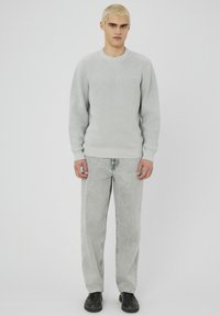 Pull en maille côtelée gris clair avec un col rond, associé à un pantalon en denim gris délavé et des chaussures noires. Le mannequin se tient devant un fond neutre.