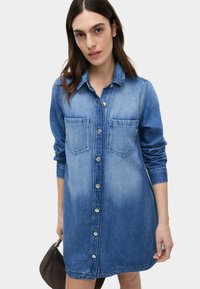 Sukienka koszulowa z denimu z dwoma kieszeniami na piersi, zapinana na guziki z przodu, z długimi rękawami oraz gradientem od ciemnoniebieskiego do jasnoniebieskiego. Miękka tekstura materiału.
